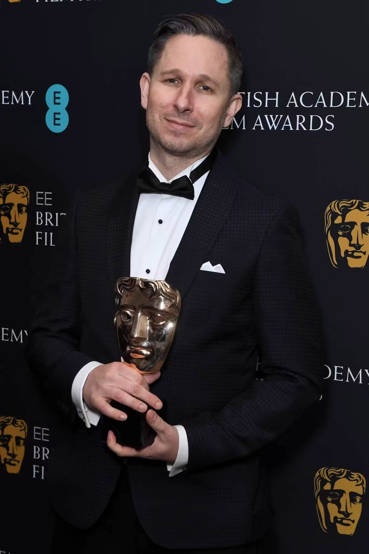 Elliot Graham in a tuxedo holding a British Academy Film Award